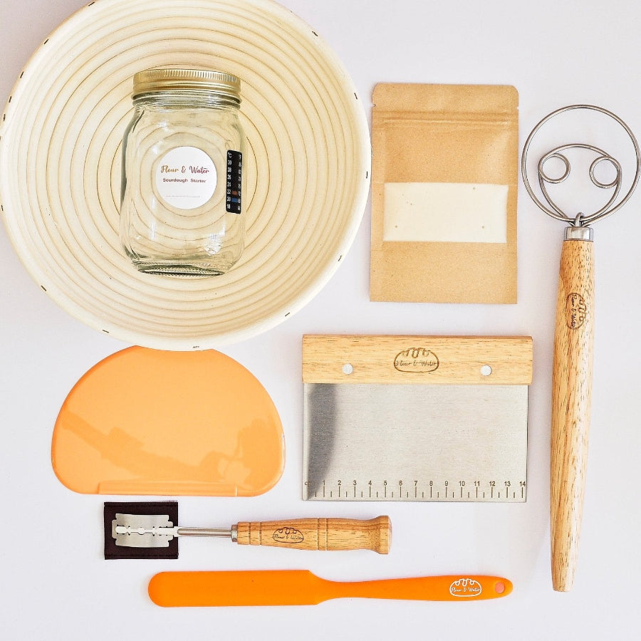 Sourdough starter kit laid out flat: rattan banneton, starter jar, dough whisk, bowl scraper, bench scraper and lame