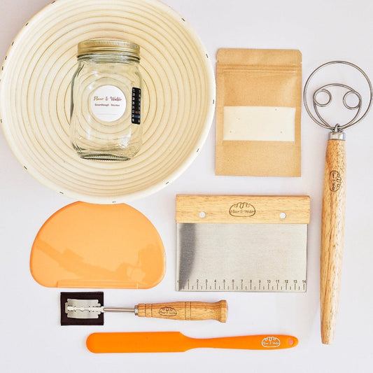Sourdough starter kit laid out flat: rattan banneton, starter jar, dough whisk, bowl scraper, bench scraper and lame